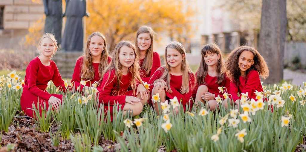 sieben Mädchen sitzen in roten Kleidern zwischen Osterglocken