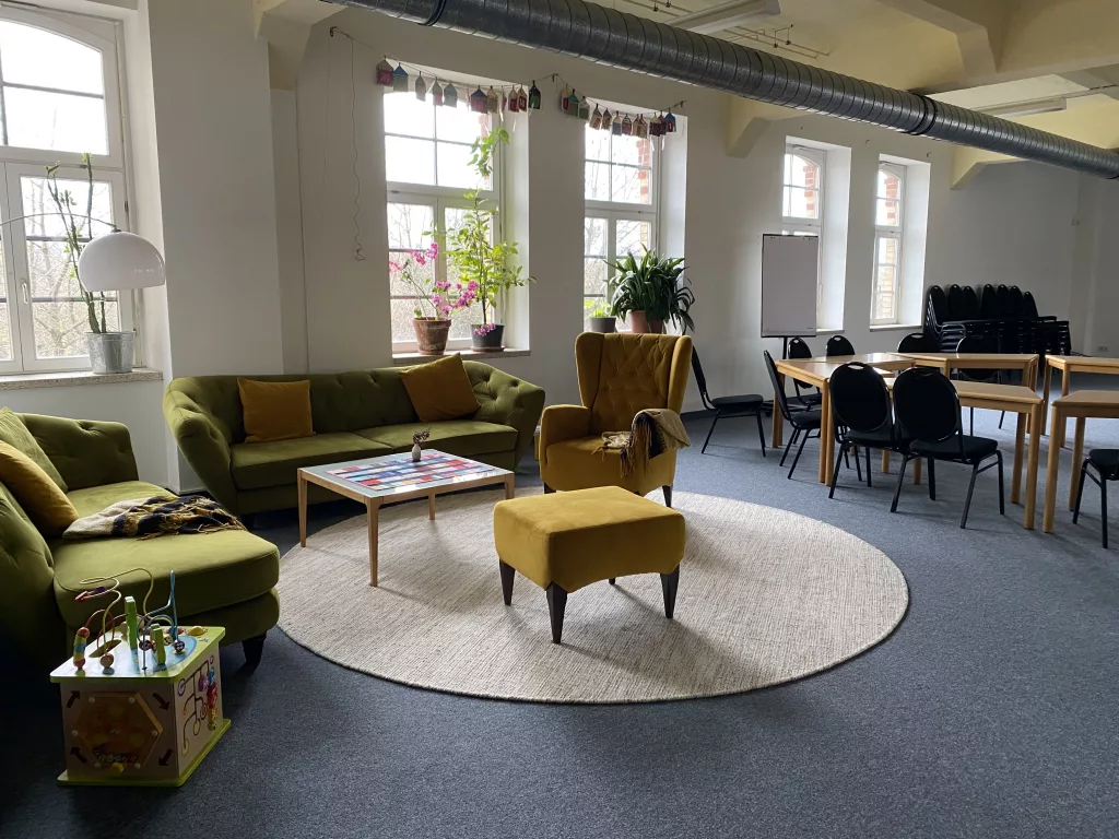 eine Sitzecke mit zwei grüne Sofas und einem gelben Sessel, in einem Saal, im Hintergrund ist noch eine Tischgruppe mit Stühlen